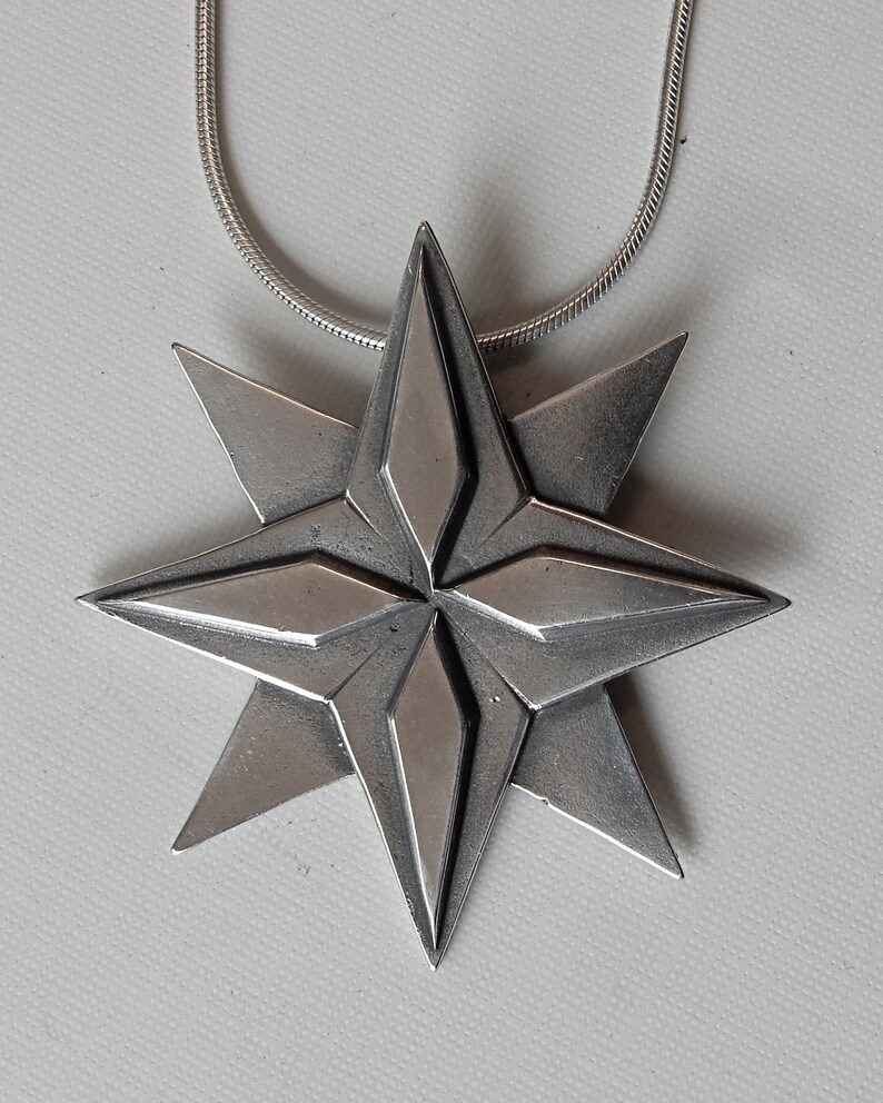 Silver coloured eight point white bronze star. Four triangular points are on the lowest layer. Four more points overlay these filling in between them.  The top four points have a raised elongated diamond shape on top of them. On chain. By Master Ark.