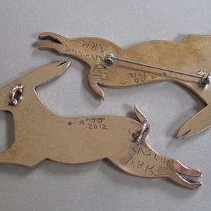 Backs of two rabbits showing two necklace loops on the back of on and a pin back on the other.  Stamped with copyright Guse 00.  Engraved with 2012, Master Ark.