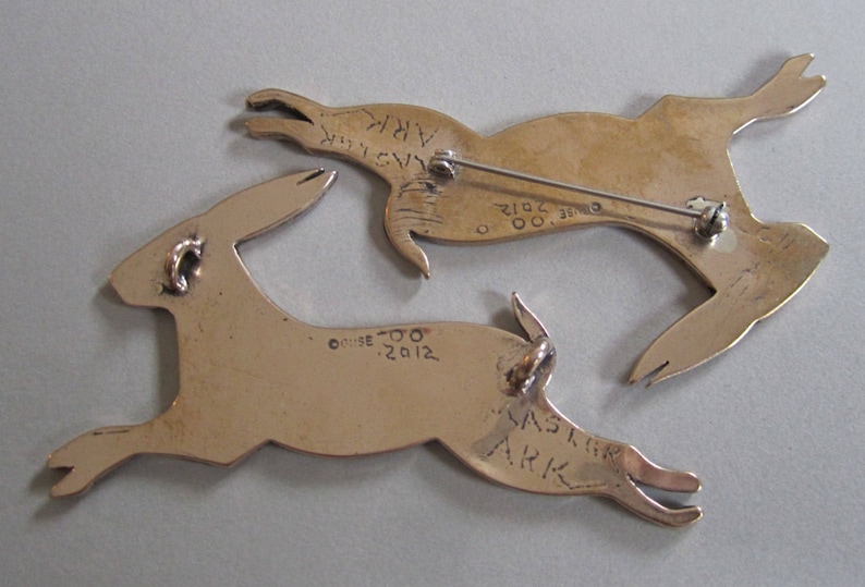 Backs of two rabbits showing two necklace loops on the back of on and a pin back on the other.  Stamped with copyright Guse 00.  Engraved with 2012, Master Ark.