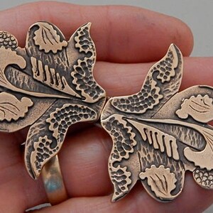 Puede incluir: Un par de broches de metal de color bronce en forma de dise&ntilde;os florales estilizados. Las broches est&aacute;n intrincadamente detalladas con patrones en espiral y tienen un acabado texturizado.