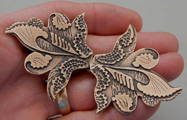 Puede incluir: Un par de broches de metal de color bronce en forma de dise&ntilde;os florales estilizados. Las broches est&aacute;n intrincadamente detalladas con patrones en espiral y tienen un acabado texturizado.