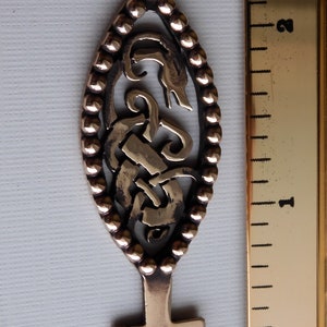 May include: A bronze key with an ornate design featuring a stylized knotwork pattern. The key has a rounded oval head and a square-shaped bow.