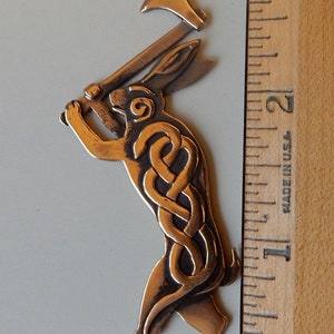 Rabbit standing on hind legs holding a broad headed axe above his head.  Body decorated with raised interlace snake pattern.  Spiral on face.  Three layers of metal.  Cast bronze by Master Ark. Shown next to ruler - three inches tall.