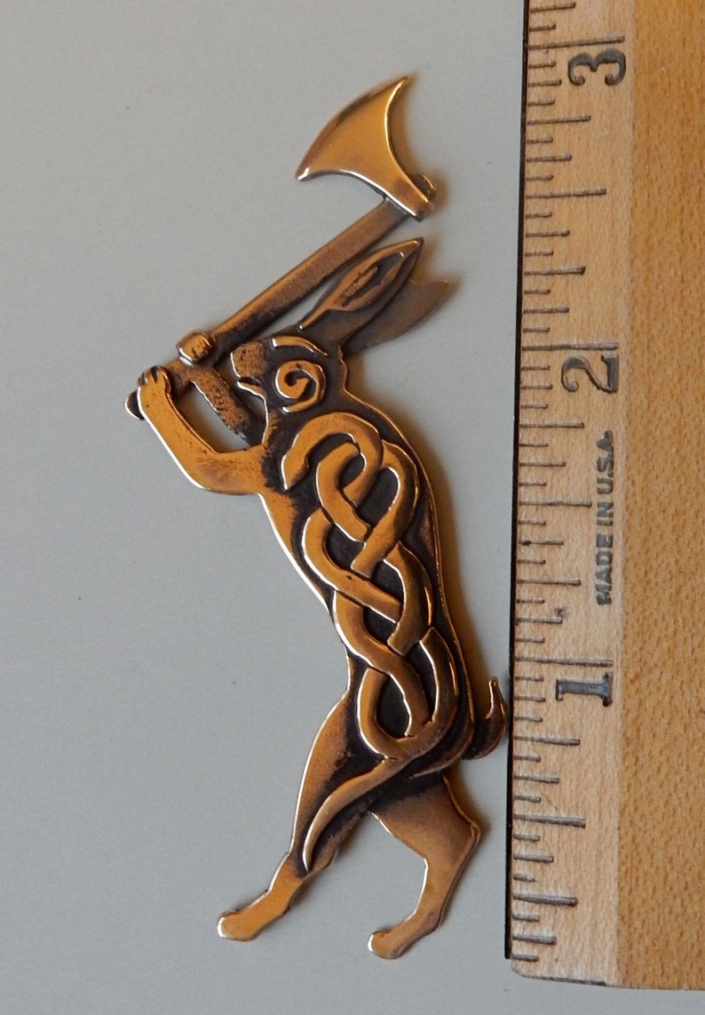 Rabbit standing on hind legs holding a broad headed axe above his head.  Body decorated with raised interlace snake pattern.  Spiral on face.  Three layers of metal.  Cast bronze by Master Ark. Shown next to ruler - three inches tall.