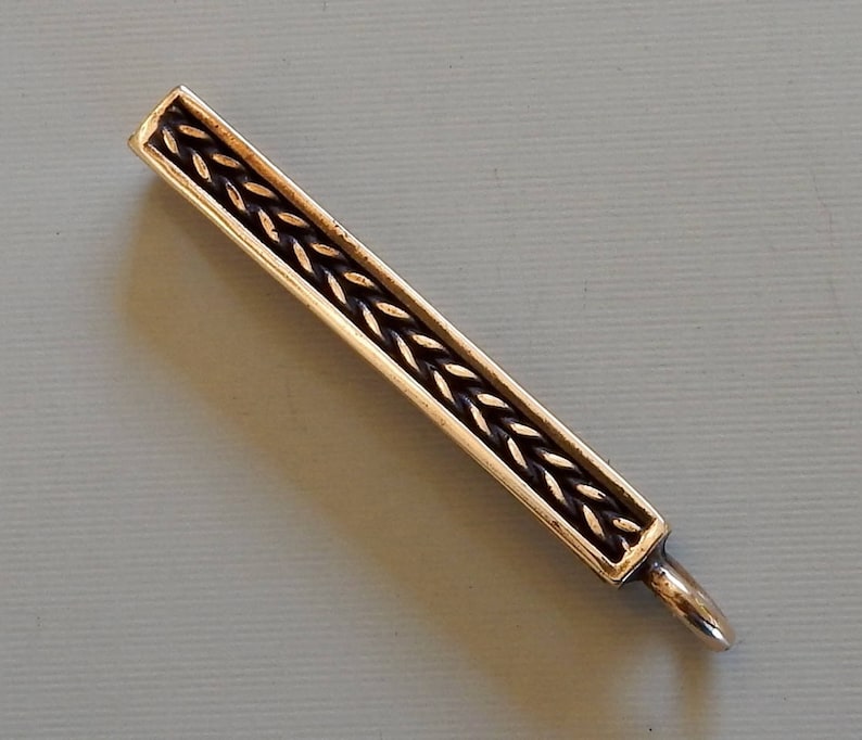 Cast bronze bar to slide onto a belt with a suspension loop at the bottom.  Herringbone wire pattern on the front in a boxed border.  By Master Ark.