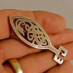 May include: A silver-toned key-shaped pendant with an intricate design of a scorpion. The pendant is oval-shaped and has a small keyhole at the top. The back of the pendant is engraved with the word "OGUIBE".