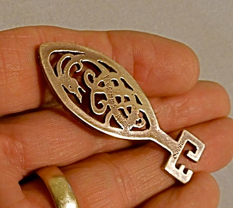 May include: A silver-toned key-shaped pendant with an intricate design of a scorpion. The pendant is oval-shaped and has a small keyhole at the top. The back of the pendant is engraved with the word "OGUIBE".