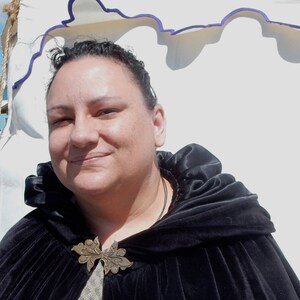 Puede incluir: Una mujer con una capa de terciopelo negro con capucha y un broche de metal decorativo. La capa tiene un cuello fruncido y se lleva sobre una camisa estampada.