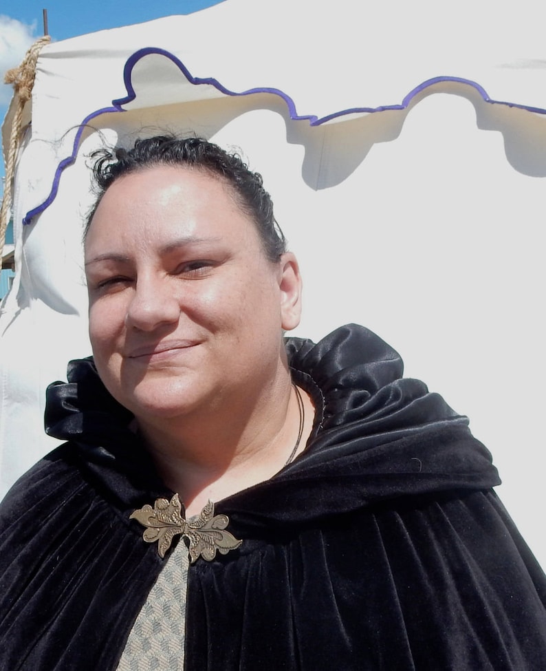 Puede incluir: Una mujer con una capa de terciopelo negro con capucha y un broche de metal decorativo. La capa tiene un cuello fruncido y se lleva sobre una camisa estampada.