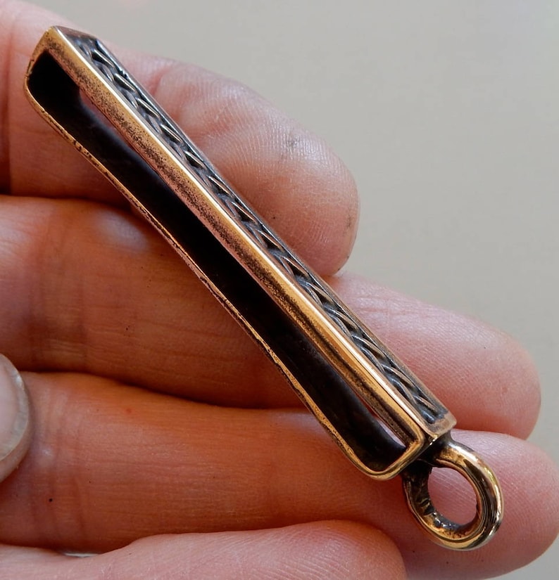 Cast bronze bar to slide onto a belt with a suspension loop at the bottom.  Herringbone wire pattern on the front in a boxed border.  3/4 view. By Master Ark.