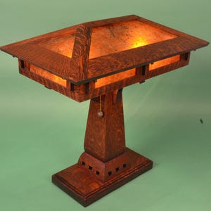 May include: A wooden table lamp with a rectangular shade and a square base. The shade is made of a translucent material and has a warm, amber glow. The lamp has a pull chain for turning it on and off.