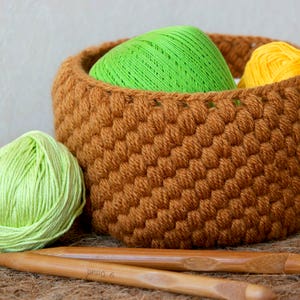 Könnte beinhalten: Ein brauner gehäkelter Korb gefüllt mit grünen und gelben Garnknäueln. Zwei Stricknadeln aus Holz liegen auf einer braunen Oberfläche.