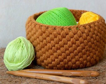 May include: A brown crocheted basket filled with green and yellow yarn balls. Two wooden knitting needles are on a brown surface.