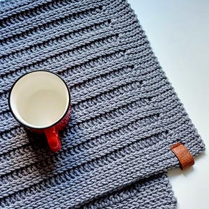 Könnte beinhalten: Ein grauer, gehäkelter Platzdeckchen mit gerippter Struktur, mit einer rot-weißen Tasse. Der Platzdeckchen hat einen Lederakzent. Die Tasse ist leer. Der Platzdeckchen ist etwa 30 cm groß.