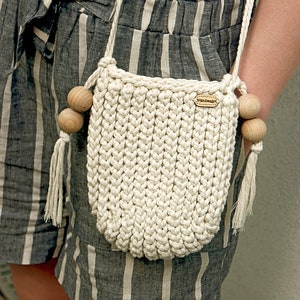 Könnte beinhalten: Cremefarbene, handgefertigte Häkeltasche mit Holzperlen und Quaste. Die Tasche hat ein gewebtes Design und ein kleines Lederetikett mit der Aufschrift "handmade". Sie wird von einem cremefarbenen Seilriemen getragen.