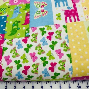 May include: A cotton fabric featuring a patchwork design of colorful panels with patterns of butterflies, giraffes, and elephants. A white ruler is visible at the bottom, showing the fabric width measures approximately 11 inches across.