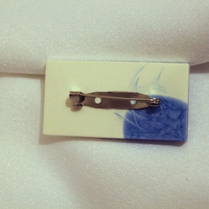 May include: A white and blue ceramic brooch with a silver pin. The brooch has a blue and white swirl design.