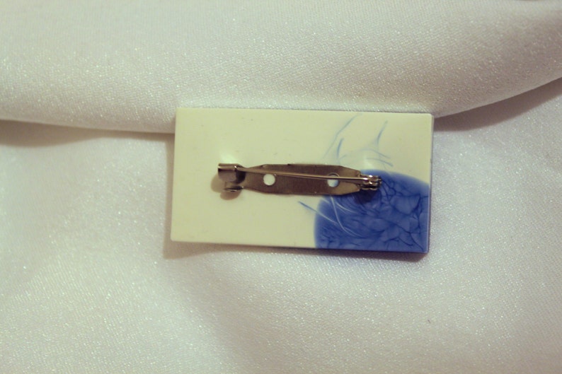 May include: A white and blue ceramic brooch with a silver pin. The brooch has a blue and white swirl design.