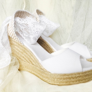 May include: White wedge sandals with a lace detail and a jute sole. The sandals are on a white tulle background.