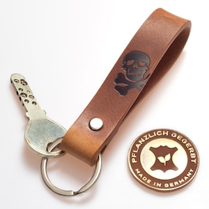 Könnte beinhalten: Ein brauner Leder-Schlüsselanhänger mit Totenkopf- und gekreuzten Knochen-Design. Der Schlüsselanhänger enthält einen silbernen Schlüsselring und einen Schlüssel. Ein rundes, goldfarbenes Emblem mit deutschem Text ist ebenfalls vorhanden.