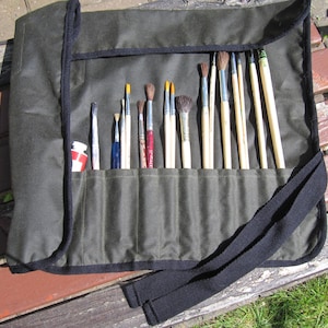 Puede incluir: Un estuche enrollable de lona verde con una correa negra y varios bolsillos para guardar pinceles. El estuche está lleno de una variedad de pinceles de diferentes tamaños y formas.