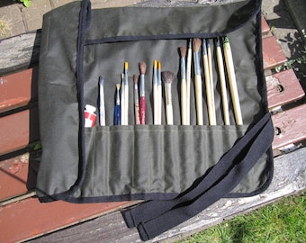 May include: A green canvas roll-up case with a black strap and multiple pockets for holding paint brushes. The case is filled with a variety of paint brushes of different sizes and shapes.