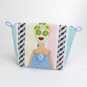 Könnte beinhalten: Eine farbenfrohe Kosmetiktasche mit Spa-Motiv. Die Tasche hat ein Streifenmuster in Pastellfarben und schwarzen Zickzacklinien. Eine Filzapplikation zeigt eine Frau mit Gurken-Augenmasken und einem blauen Kleid.