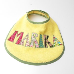 Könnte beinhalten: Ein gelber Frottee-Babyspucktuch mit grünem Rand und dem Namen "MARIKA" in bunten Stoffbuchstaben.