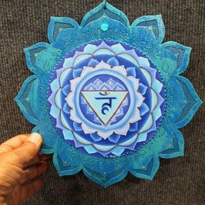 May include: A blue and gold mandala with a white triangle in the center. The triangle contains the Sanskrit symbol for the fifth chakra, Vishuddha, which is the throat chakra. The mandala is painted on a wooden surface and has a teal blue border.