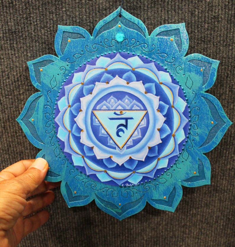 May include: A blue and gold mandala with a white triangle in the center. The triangle contains the Sanskrit symbol for the fifth chakra, Vishuddha, which is the throat chakra. The mandala is painted on a wooden surface and has a teal blue border.