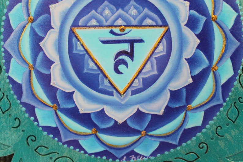 May include: A blue and gold mandala with a turquoise triangle and Sanskrit symbol in the center. The mandala is painted on a wooden surface.