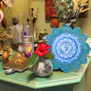 May include: A blue and turquoise mandala with the symbol "5" in the center. The mandala is surrounded by a decorative border. The mandala is on a green surface with a small vase and a red flower.