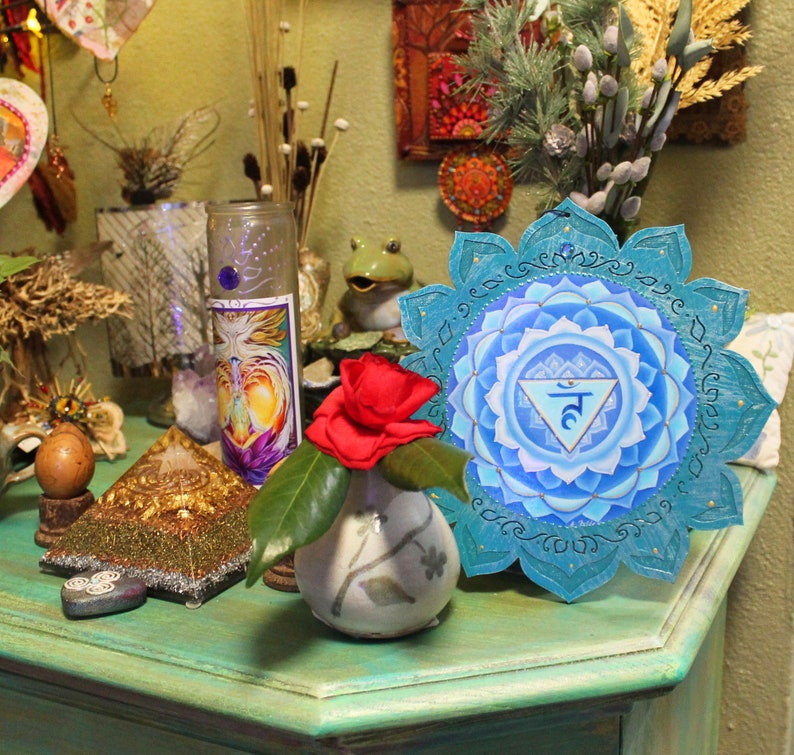 May include: A blue and turquoise mandala with the symbol "5" in the center. The mandala is surrounded by a decorative border. The mandala is on a green surface with a small vase and a red flower.