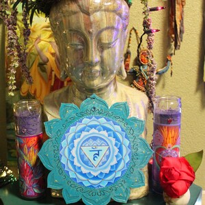 May include: A teal blue mandala with a white and blue inner circle and a gold symbol in the center. The mandala is surrounded by a white border with a gold trim. The mandala is hanging on a wall with a statue of Buddha in the background.