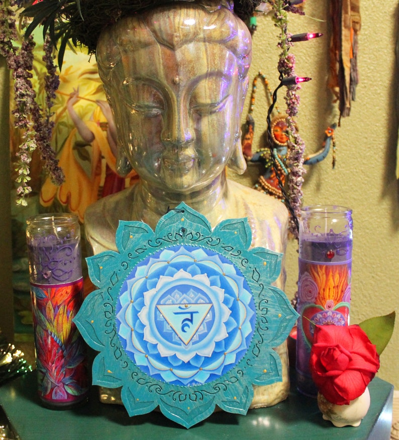 May include: A teal blue mandala with a white and blue inner circle and a gold symbol in the center. The mandala is surrounded by a white border with a gold trim. The mandala is hanging on a wall with a statue of Buddha in the background.
