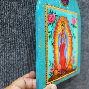 May include: A hand-painted, turquoise-colored religious icon featuring a depiction of the Virgin Mary. The image is framed with gold trim and adorned with pink roses and gold accents. The icon is held against a gray background.