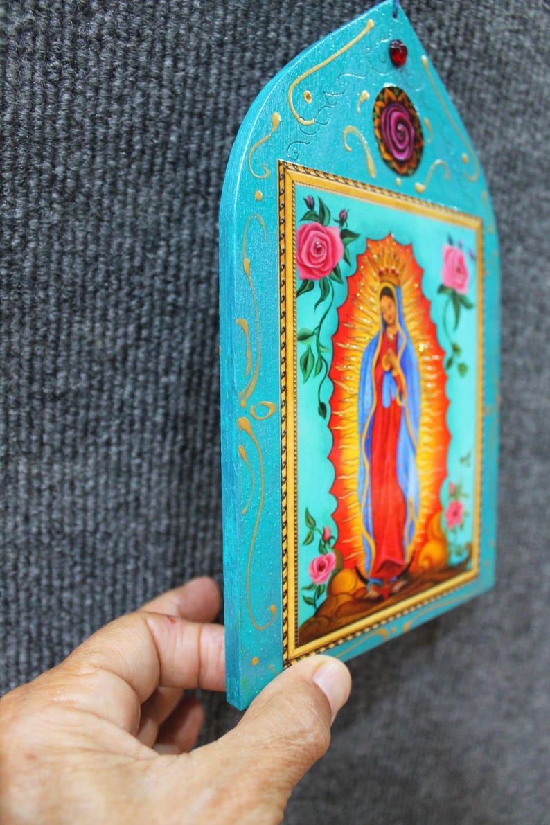 May include: A hand-painted, turquoise-colored religious icon featuring a depiction of the Virgin Mary. The image is framed with gold trim and adorned with pink roses and gold accents. The icon is held against a gray background.