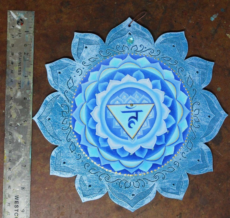 May include: A blue and white mandala with a gold trim and a blue triangle with the Sanskrit symbol "Ham" in the center. The mandala is painted on a wooden surface and has a distressed look.