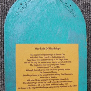 May include: A turquoise wooden plaque with a decorative top and a rectangular yellow insert. The insert features text about Our Lady of Guadalupe. The plaque has a small hole at the top for hanging.