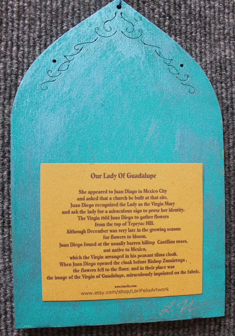 May include: A turquoise wooden plaque with a decorative top and a rectangular yellow insert. The insert features text about Our Lady of Guadalupe. The plaque has a small hole at the top for hanging.