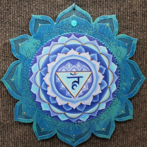 May include: A blue and gold mandala with a turquoise border and a blue triangle with the Sanskrit symbol "Ham" in the center. The mandala is painted on a wooden surface.