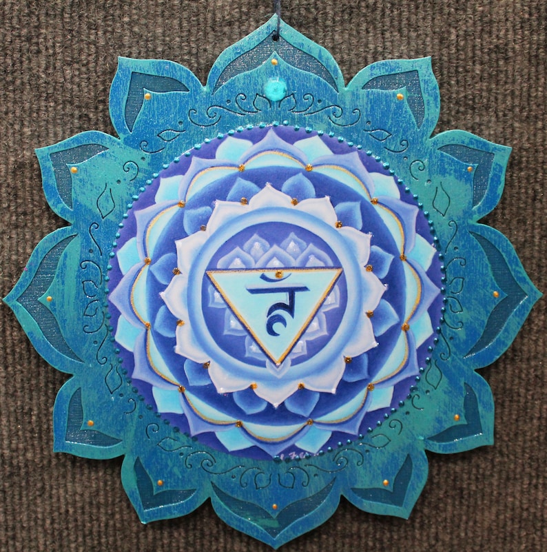 May include: A blue and gold mandala with a turquoise border and a blue triangle with the Sanskrit symbol "Ham" in the center. The mandala is painted on a wooden surface.