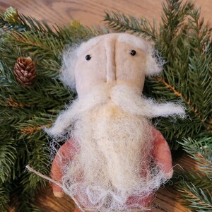 Könnte beinhalten: Eine handgemachte Weihnachtsmann-Schrankpuppe mit weißem Bart und roter Stoffkleidung. Die Puppe ist von grünen Kiefernästen umgeben. Der Text "The Old Dusty Cupboard copy right 2023 Father Christmas cupboard doll" ist unter der Puppe gedruckt.