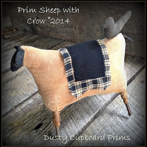 Puede incluir: Un cordero de tela beige con una manta de cuadros negros y blancos y un cuervo negro posado en su espalda. El cordero tiene patas de alambre oxidado y está sentado sobre una superficie de madera. El texto "Prim Sheep with Crow 2014" está escrito encima del cordero. El texto "Dusty Cupboard Prims" está escrito debajo del cordero.