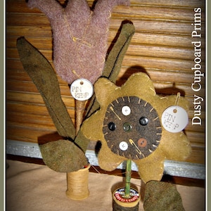 May include: Two felt flower pinkeeps, one a brown flower with a brown center and the other a purple flower. Both flowers have a pinkeep sign and are sitting on top of spools of thread.