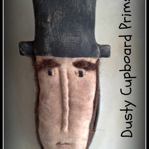 Puede incluir: Una muñeca de fieltro hecha a mano de Abraham Lincoln con un sombrero de copa negro. La muñeca está etiquetada como "Dusty Cupboard Prims" y "Abe Lincoln".