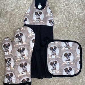 May include: A set of kitchen towels and pot holders featuring a brown and white boxer dog pattern. The towels and pot holders have black trim and the word "Boxer" is repeated throughout the pattern.