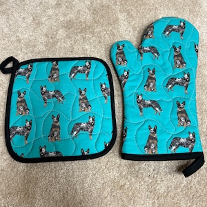 May include: A set of two teal blue oven gloves and a pot holder with a black trim. The gloves and pot holder feature a repeating pattern of Australian Shepherd dogs.