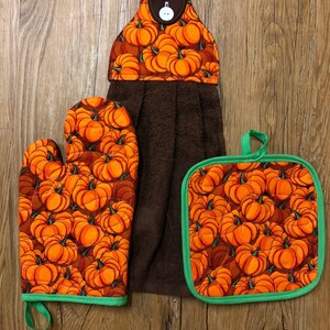 Puede incluir: Un juego de tres accesorios de cocina con estampado de calabaza marrón y verde. El juego incluye una toalla de cocina marrón con un lazo para colgar, un guante de horno verde y un posavasos verde.
