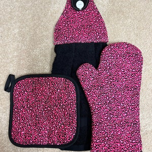 May include: A pink and black kitchen towel set with a leopard print design. The set includes a potholder, a hot mitt, and a hanging towel.